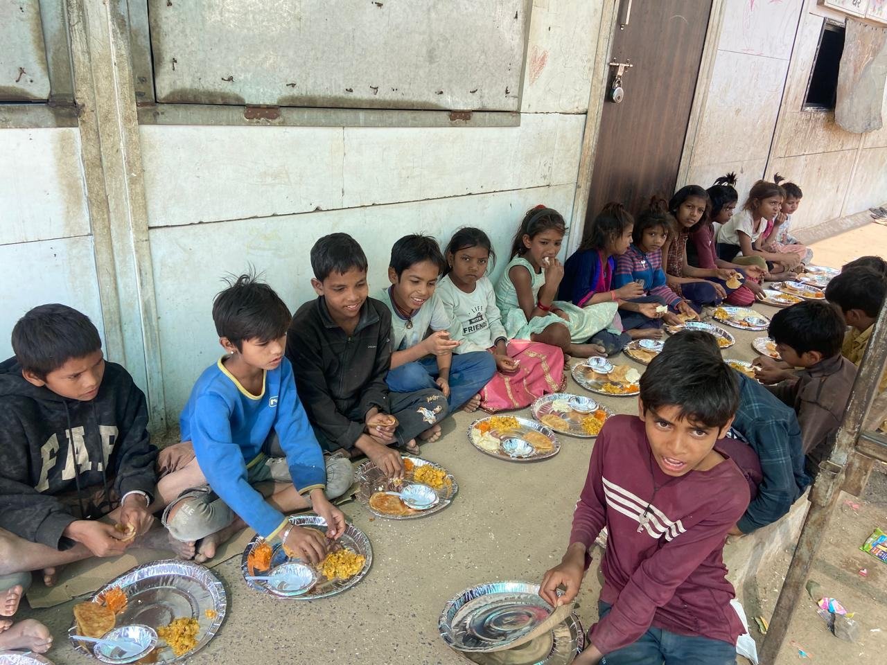 Children seated in a circle with steel plates of hot food